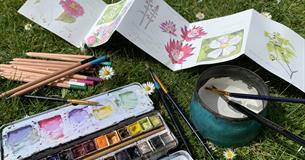 Aphotograph of a concetrina of water coulor paperwith Victorian style flowers painted on it,. The paper is surrounded by a box of watercolour paints,