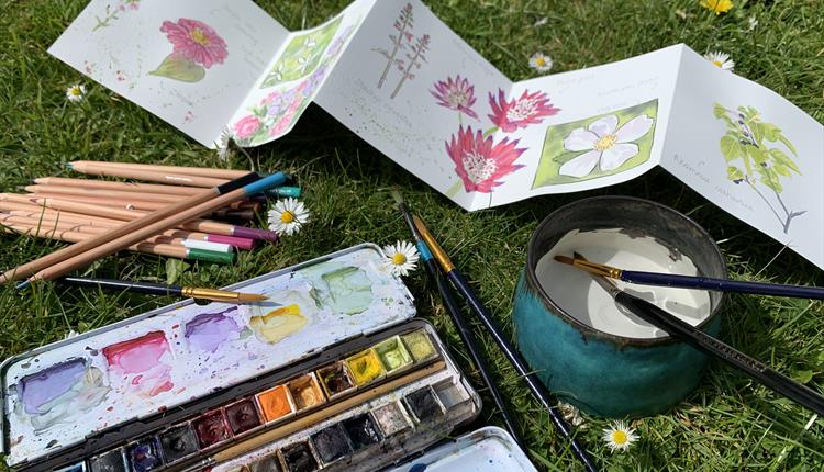 Aphotograph of a concetrina of water coulor paperwith Victorian style flowers painted on it,. The paper is surrounded by a box of watercolour paints,
