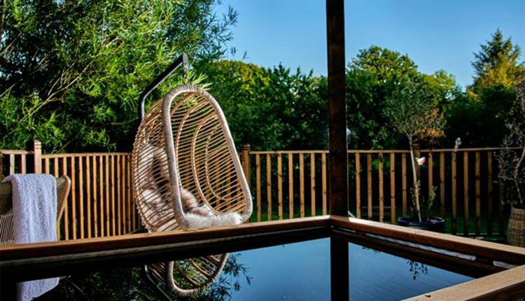 A wicker hanging chair with a cushion is on a wooden deck beside a fenced yard with trees and plants; water is visible in the foreground.