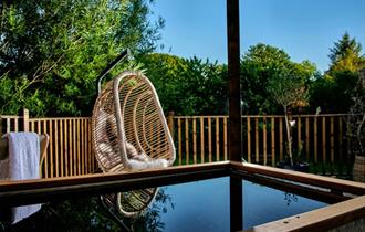 A wicker hanging chair with a cushion is on a wooden deck beside a fenced yard with trees and plants; water is visible in the foreground.