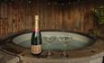A bottle of Montaudon champagne and two elegant glasses sit beside a steaming hot tub, framed by rustic wooden walls.
