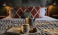 A cosy scene featuring a cup labeled "You, Me & Coffee," a teapot, snacks, and a book on a plush blanket in a softly lit bedroom.