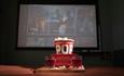 A cinema screen behind a red tub of popcorn and two drinks.