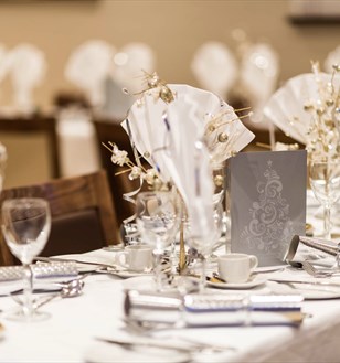 Elegant table setting with white napkins folded in glasses, ornate centrepieces, and silver crackers on a white tablecloth, conveying a festive atmosphere.