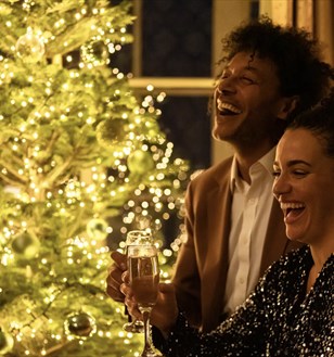 A joyful couple in festive attire holds champagne glasses, laughing beside a beautifully decorated, glowing Christmas tree. The scene conveys warmth and celebration.