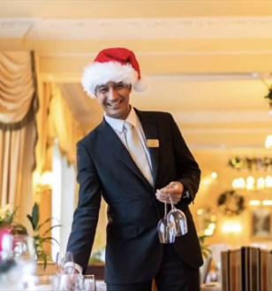 A smiling waiter in a suit and Santa hat holds two wine glasses in a festive, warmly lit restaurant adorned with garlands and elegant decor.