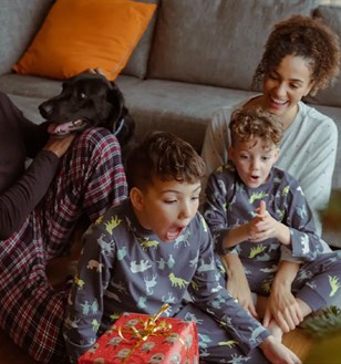 Family in pyjamas sits on the floor near a Christmas tree, excitedly opening gifts. A dog joins, creating a joyful, festive atmosphere.