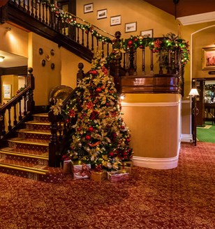 Festive hotel lobby with a decorated Christmas tree beside ornate wooden stairs. Warm lighting and garlands create a cosy, inviting holiday ambience.